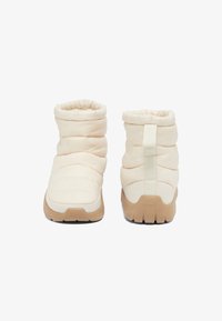 Botas de tobillo de color crema y con forma acolchada, de tela texturizada, con elástico en la parte superior y suela de goma marrón. Con un tirador en la parte trasera para facilitar su uso.