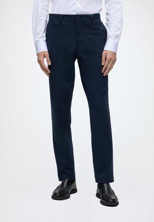 Homme portant un pantalon de costume bleu marine, des chaussures habillées noires et une chemise blanche à manches longues boutonnée, debout devant un fond neutre.