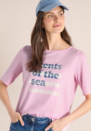 Femme portant une casquette bleu clair, un t-shirt rose avec le texte "Scents of the sea" et un jean bleu, regardant sur le côté les mains dans les poches.