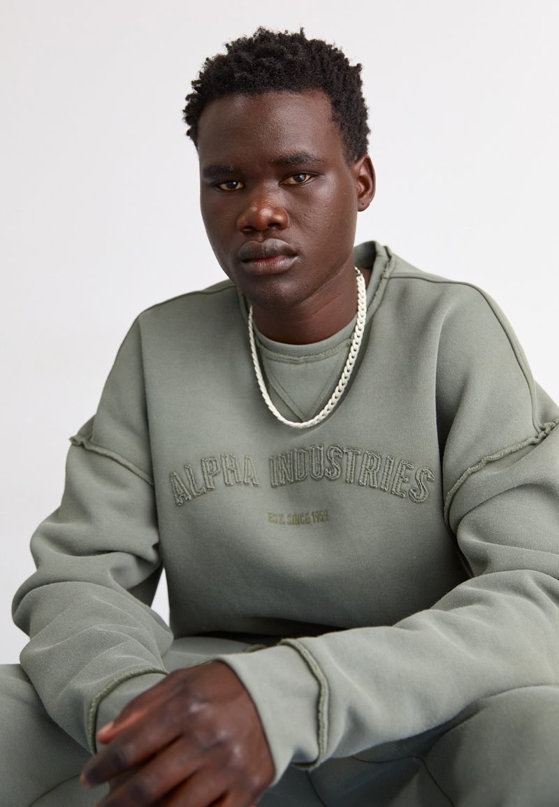 Grau-grüner Sweatshirt mit "ALPHA INDUSTRIES" Stickerei, lockere Passform, Raglanärmel, weiche Textur, begleitet von einer silbernen Kettenkette.