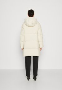 Manteau bouffant couleur crème avec capuche, présentant un design matelassé et des manches longues. Porté par-dessus un pantalon sombre et des chaussures à talons.
