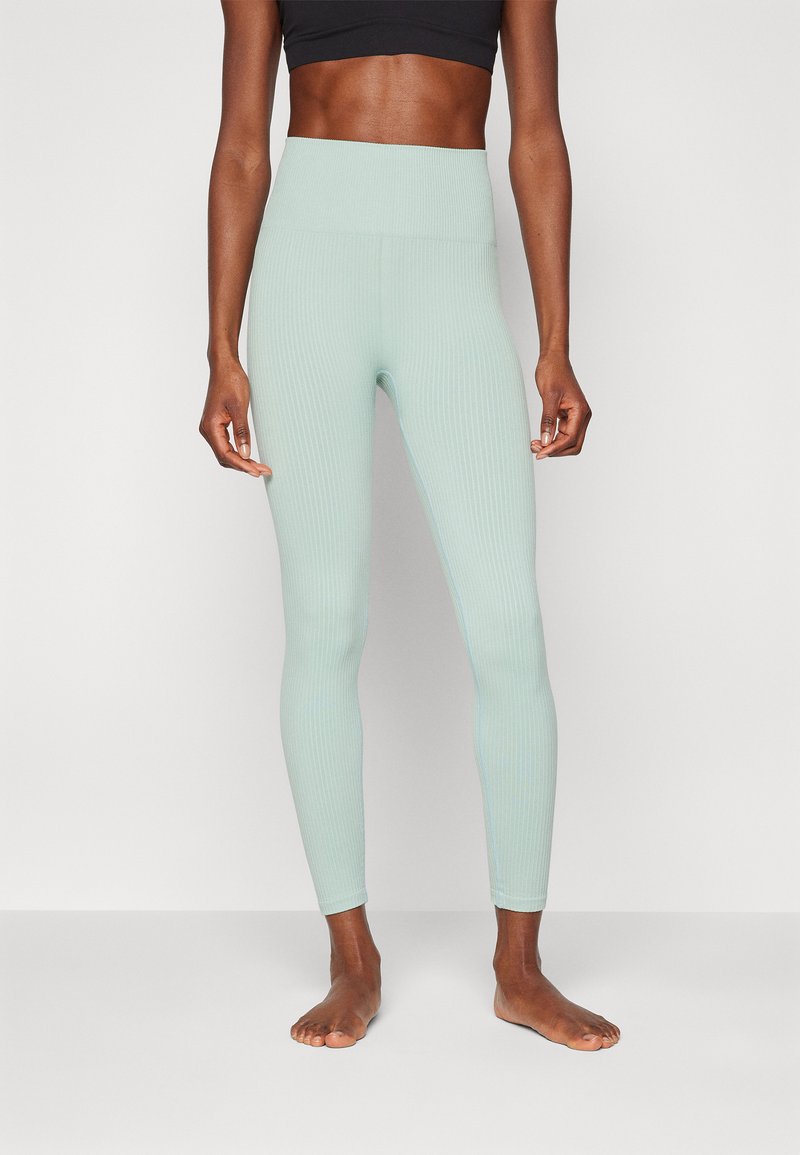 Leggings a coste di color verde chiaro con vita alta, texture liscia e design aderente, esibiti senza scarpe su uno sfondo neutro.