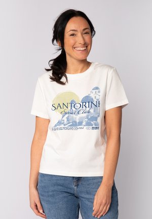 Frau mit dunklen Haaren im Pferdeschwanz lächelt, trägt ein weißes T-Shirt mit dem Aufdruck "Santorini Sunset Club" und blaue Jeans vor schlichtem Hintergrund.