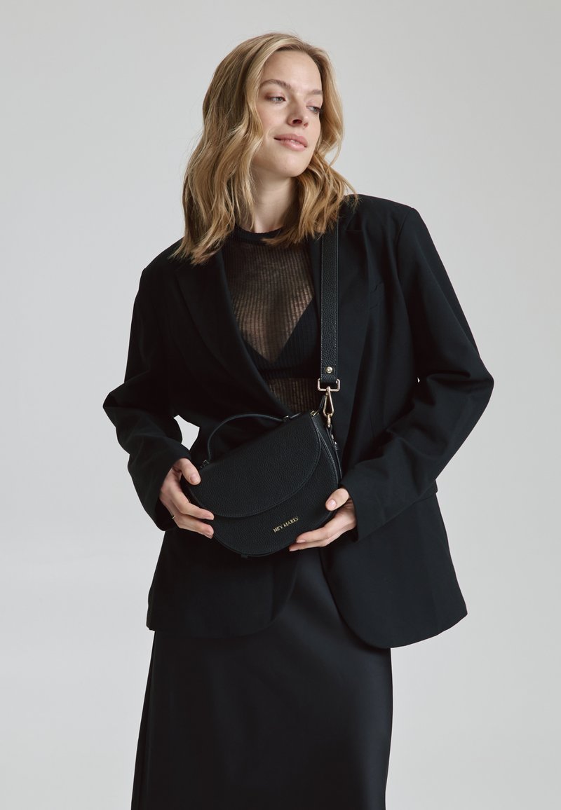 Zwarte leren tas met een gebogen silhouet, textuur oppervlak en gouden details. Draagt bij een zwarte blazer en jurk, die gelaagde ontwerpen toont.