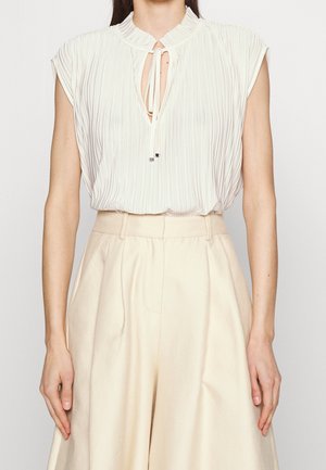 Blouse blanche sans manches plissée avec un col à nouer, rentrée dans un pantalon large crème taille haute portée par une personne.