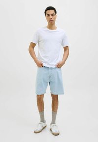 Camiseta de algodón blanca, pantalones cortos de mezclilla azul claro, calcetines grises y zapatillas blancas con acentos en color marrón. El modelo está de pie con las manos en los bolsillos.