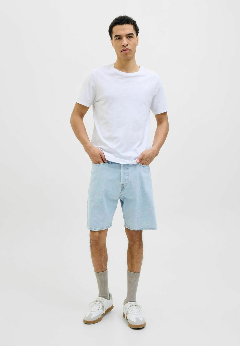 Camiseta de algodón blanca, pantalones cortos de mezclilla azul claro, calcetines grises y zapatillas blancas con acentos en color marrón. El modelo está de pie con las manos en los bolsillos.