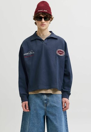 Noor mees kannab tumesinist plekiga dressipluusi, punast mütsi, tumedaid päikeseprille, beeži aluspluusi ja lahtisi siniseid teksapükse ühevärvilise tausta ees.