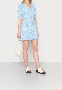 Kurzes blaues Blumenkleid mit Puffärmeln, V-Ausschnitt und schwingendem Rock. Kombiniert mit weißen Riemchensandalen und einer weißen strukturierten Handtasche.
