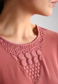 Rosa Bluse aus texturiertem Stoff mit einem Spitzenbesatz entlang des Ausschnitts, der ein dreieckiges Design mit offenen und geschlossenen Gewebemustern schafft.