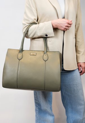 Groene leren tote bag met dubbele handgrepen, gladde textuur en gestikte accenten; vastgehouden door een persoon in een beige blazer en blauwe jeans.