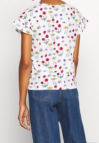 Haut à manches courtes, blanc avec des fleurs rouges, jaunes et violettes, présentant des manches à volants froncées et une coupe décontractée. Porté avec un jean bleu.