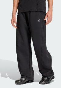 Černé tepláky s volnějším střihem, vyrobené z měkkého materiálu, s jemnými švovými detaily a logem Adidas ve stříbrné reflexní barvě.