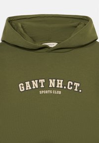 Maslinasto zeleni dukserica napravljena od mekog pamuka. Sadrži veliki printani logo "GANT NH. CT. SPORTS CLUB" u boji slonovače i tamnim akcentima.