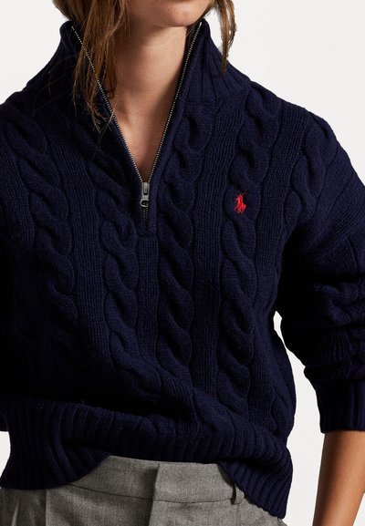 Woman wearing a navy blue cable-knit zip-up sweater with a red horse logo and gray pants, shown from neck to waist.