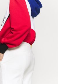 Person in weißen Hosen und einer rot, weiß und blauen Jacke mit Farbblockierung, die mit einer Hand in der Tasche an einem schlichten Hintergrund steht.