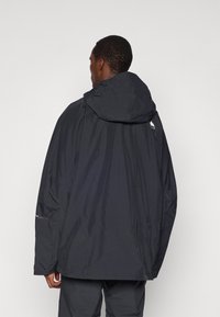 Chaqueta impermeable negra con capucha, con un ajuste holgado, puños elásticos y un logo en la parte posterior. Fabricada con un material ligero y duradero.