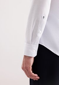 Mano di un uomo al lato, che indossa una camicia a scacchi bianca con pantaloni neri; sulla manica è ricamato un piccolo rose nero vicino ai bottoni del polsino.