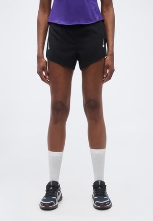 Osoba nosiaca čierne bežecké šortky Nike, fialové tričko, biele ponožky a čierne športové topánky stojí na jednofarebnom pozadí.