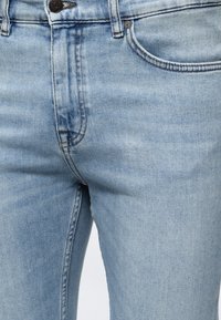 Világoskék farmernadrágok elölről, közeli kép, amely gombot, cipzárt és az elülső zseb varrásának részleteit mutatja.
