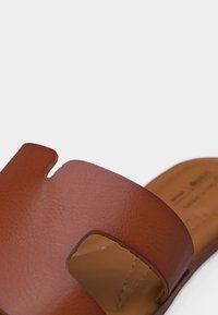 Sandale marron en cuir à glissière avec une large sangle présentant un design découpé en forme de H sur un fond clair.