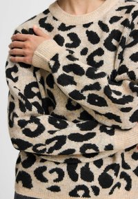 Pull beige à imprimé léopard noir. Col et poignets côtelés. Tissu doux et texturé avec des manches longues. Le motif présente des formes variées.