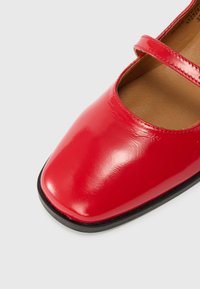 Červená lakovaná Mary Jane bota s černou podrážkou a jedním popruhem na bílém pozadí, ukazující špičku a část stélky.