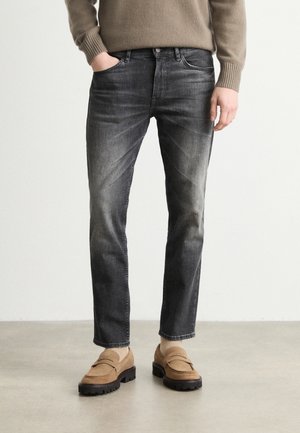 Man draagt vervaagde zwarte slim-fit jeans, beige trui en lichtbruine suède loafers met sokken, staand tegen een egaal lichte achtergrond.