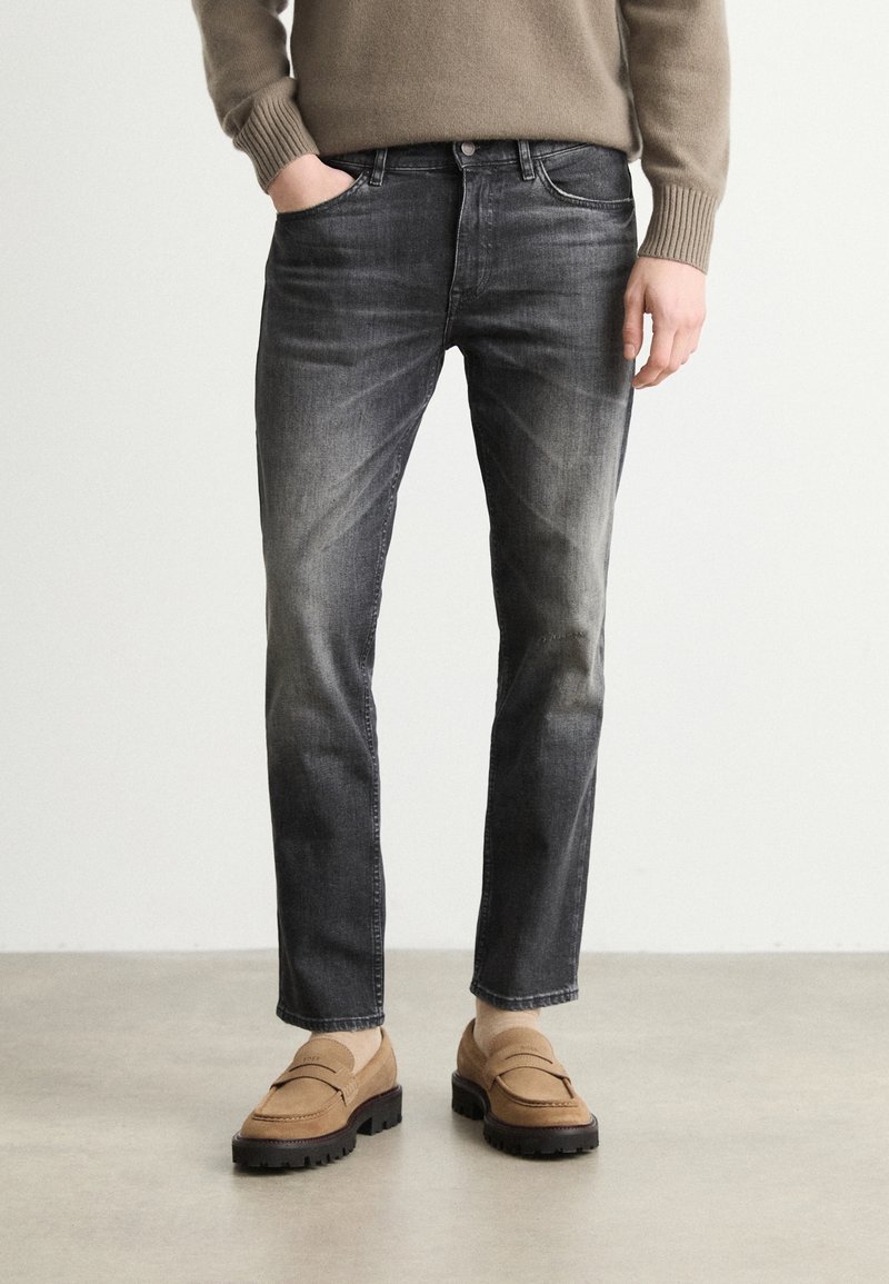 Homme portant un jean noir slim délavé, un pull beige et des mocassins en daim marron clair avec des chaussettes, debout devant un fond clair uni.