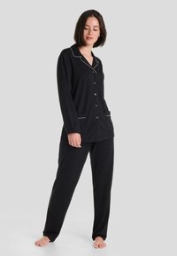 Conjunto de pijama negro que incluye una camisa de botones con ribetes blancos en contraste y bolsillos laterales, acompañada de pantalones holgados. Tela suave, ajuste relajado.
