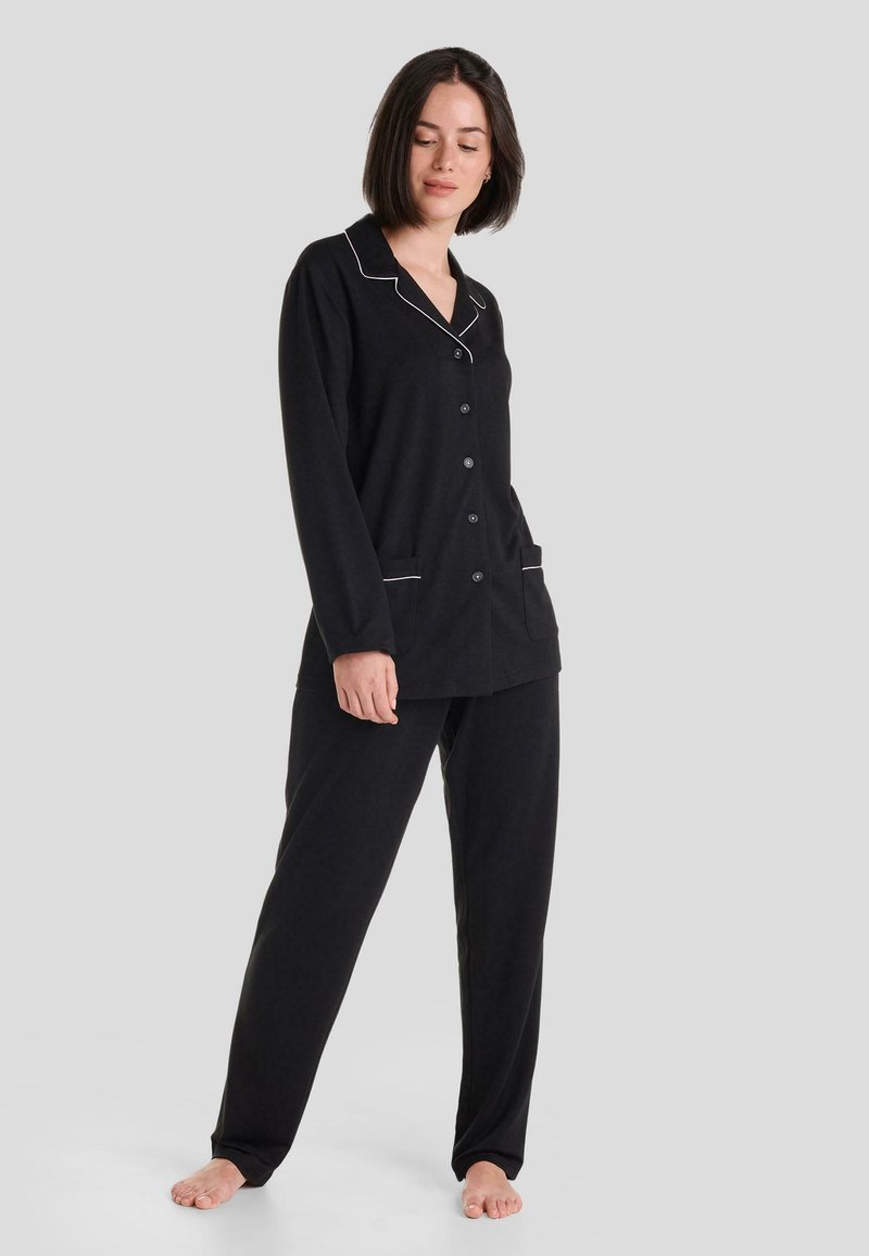 Conjunto de pijama negro que incluye una camisa de botones con ribetes blancos en contraste y bolsillos laterales, acompañada de pantalones holgados. Tela suave, ajuste relajado.