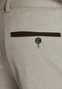 Pantaloni din țesătură bej în model herringbone, cu o margine de catifea maro pe buzunarul din spate, având un sistem de închidere cu patru butoane negre.