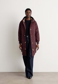 Bordeaux gewatteerde lange jas met een capuchon, voorzien van gouden knopen en een contrasterende voering, gedragen over een zwarte top en blauwe denim broek.