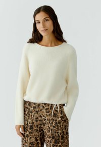 Vrouw met lang, donker haar, gekleed in een zachte witte sweater en bruine kleding met luipaardprint, met haar rechterhand in de zak op een lichte achtergrond.