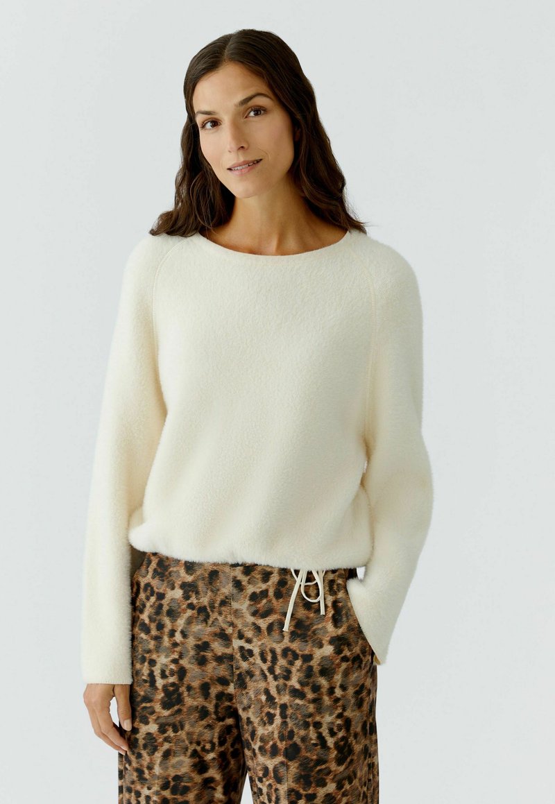 Vrouw met lang, donker haar, gekleed in een zachte witte sweater en bruine kleding met luipaardprint, met haar rechterhand in de zak op een lichte achtergrond.