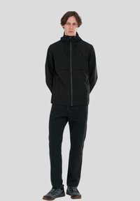 Sort lynlåsjakke med hætte, der har en frontlomme og elastiske manchetter, parret med sorte jeans og grå sko. Ensfarvet baggrund.