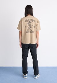 Beige cotton T-shirt with graphic print on back, featuring a cartoon figure and text "DICKIES LAND AHOY!!" in black. Paired with black pants.