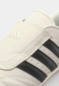 Svetlo sivi teniski z brezskrbno teksturo, črne usnjene akcentne črte in embosirano Adidasovo logo na jeziku. Oblikovanje z ravnim podplatom.