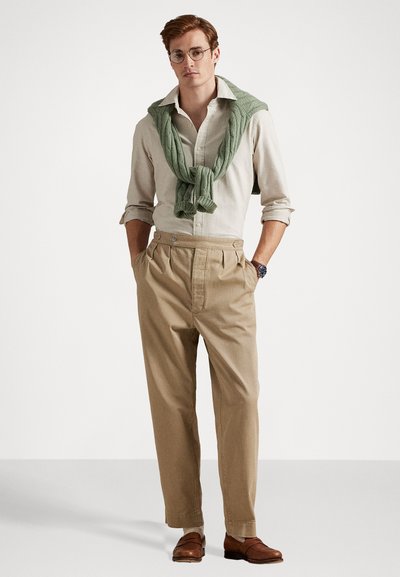 Beige trousers with pleats, light button-up shirt, and green knit sweater tied at the neck. Brown leather shoes and round glasses complete the look.