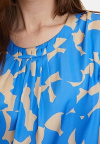 Närbild av en kvinnas halslinje som bär en blå blus med beige abstrakta blommönster och små plisseringar nära den runda kragen.