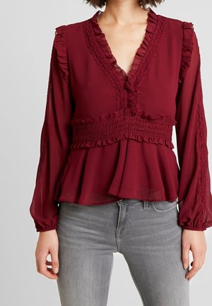 Kvinde iført bordeaux rød bluse med lange ærmer, flæser, blondekanter, rynket talje og lysegrå tætsiddende jeans mod en ensfarvet baggrund.