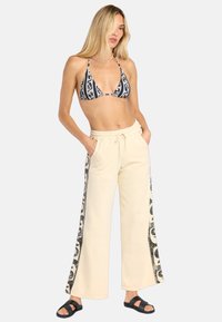 Top de bikini de patrón en blanco y negro con copas en triángulo, combinado con pantalones de chándal de pierna ancha en beige claro con acentos laterales en negro. Sandalias negras.