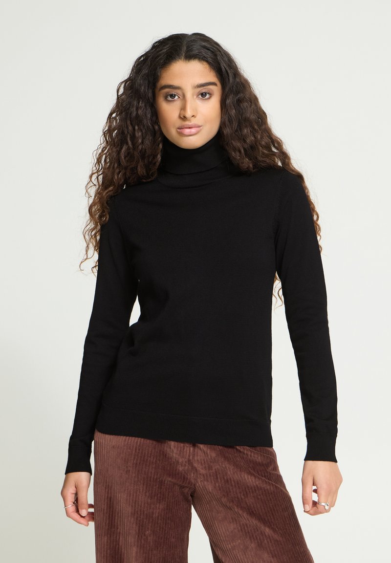 Femme aux longs cheveux bouclés, portant un pull à col roulé noir et un pantalon en velours côtelé marron, debout sur un fond uni.