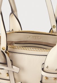 Interior de uma bolsa de pele creme com fecho dourado, apresentando um bolso e um logótipo da marca em relevo. Realçada com bordas pretas e ferragens douradas.