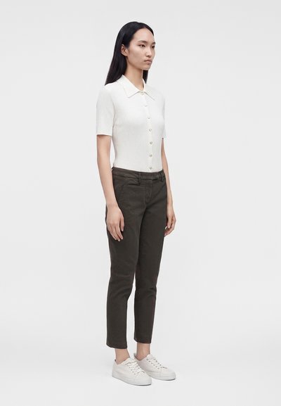 Femme aux longs cheveux noirs portant un haut blanc à manches courtes boutonné, un pantalon foncé et des baskets blanches, debout devant un fond uni.