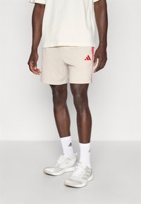 Μπεζ αθλητικό σορτς με κόκκινες πλευρικές ρίγες και λογότυπο Adidas. Φορεμένο με λευκές κάλτσες και ανοιχτό γκρι �αθλητικά παπούτσια.