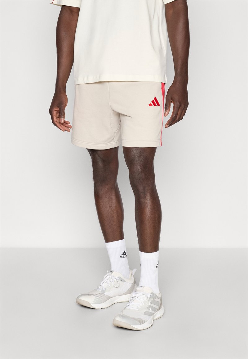 Μπεζ αθλητικό σορτς με κόκκινες πλευρικές ρίγες και λογότυπο Adidas. Φορεμένο με λευκές κάλτσες και ανοιχτό γκρι αθλητικά παπούτσια.