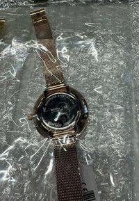 Ronde horloge van roestvrij staal met een zwarte wijzerplaat, een metalen mesh-band, een datumweergave en gegraveerde details. Waterbestendig ontwerp.