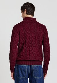 Bordeauxfarbener Kabelstrickpullover mit hohem Kragen und geripptem Saum, der ein strukturiertes Muster und taillierte Ärmel aufweist. Kombiniert mit blauen Jeans.