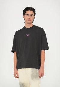 Camiseta de gran tamaño en gris oscuro, de cuello redondo y mangas cortas. Presenta un pequeño logo rosa de "Reebok" en el pecho. Material de algodón suave.